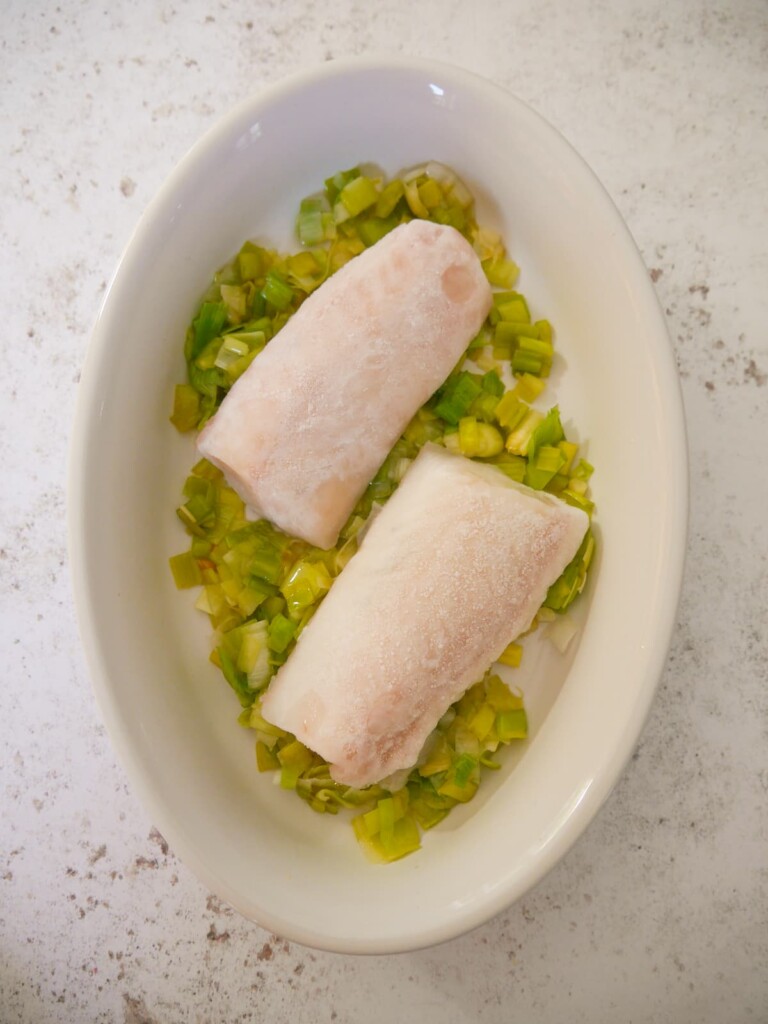 A white oven proof dish with 2 frozen cod fillets on top of a bed of sauteed leeks.