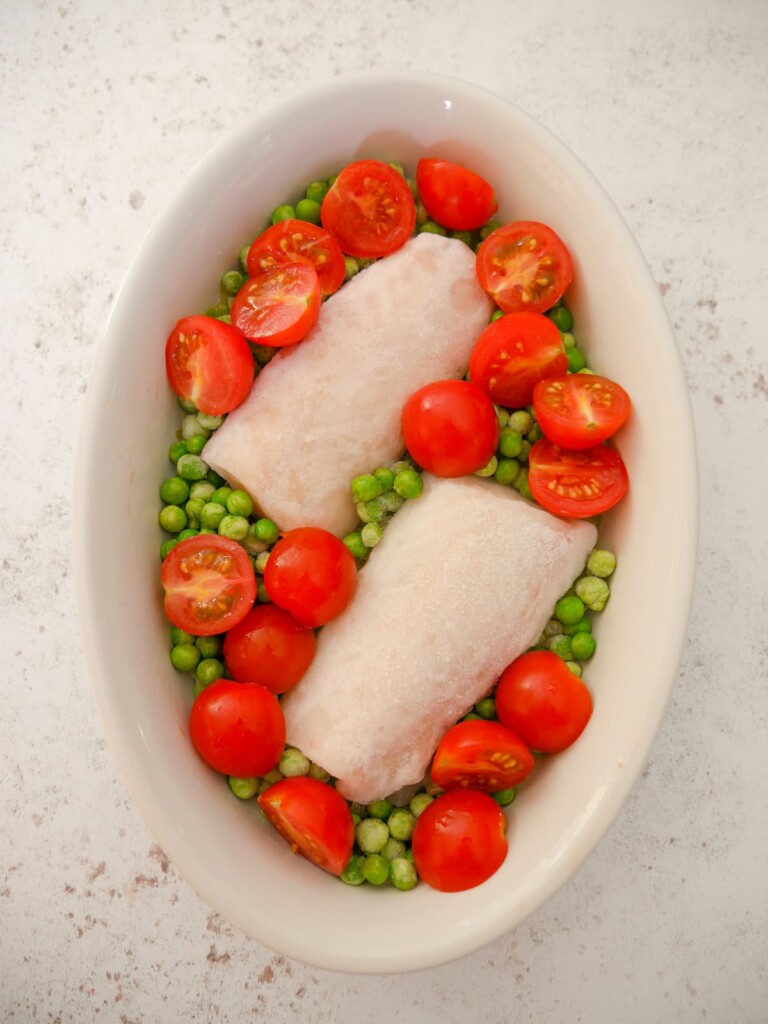 A white oven proof dish filled with cod fillets, garden peas and cherry tomato halves.