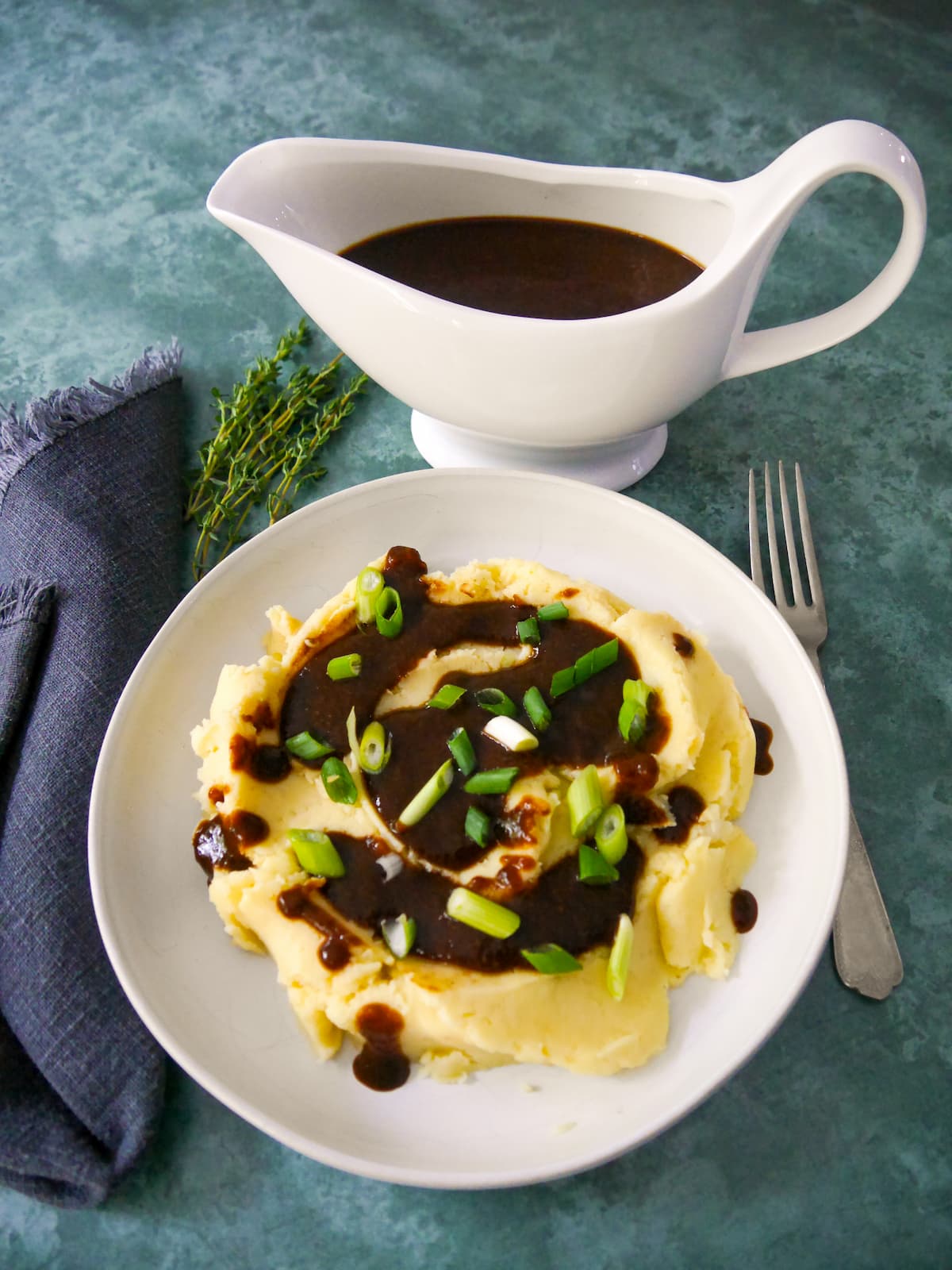 A bowl of creamy mashed potatoes topped with vegan gravy and garnished with sliced green onions, with a gravy boat of gravy set alongside.