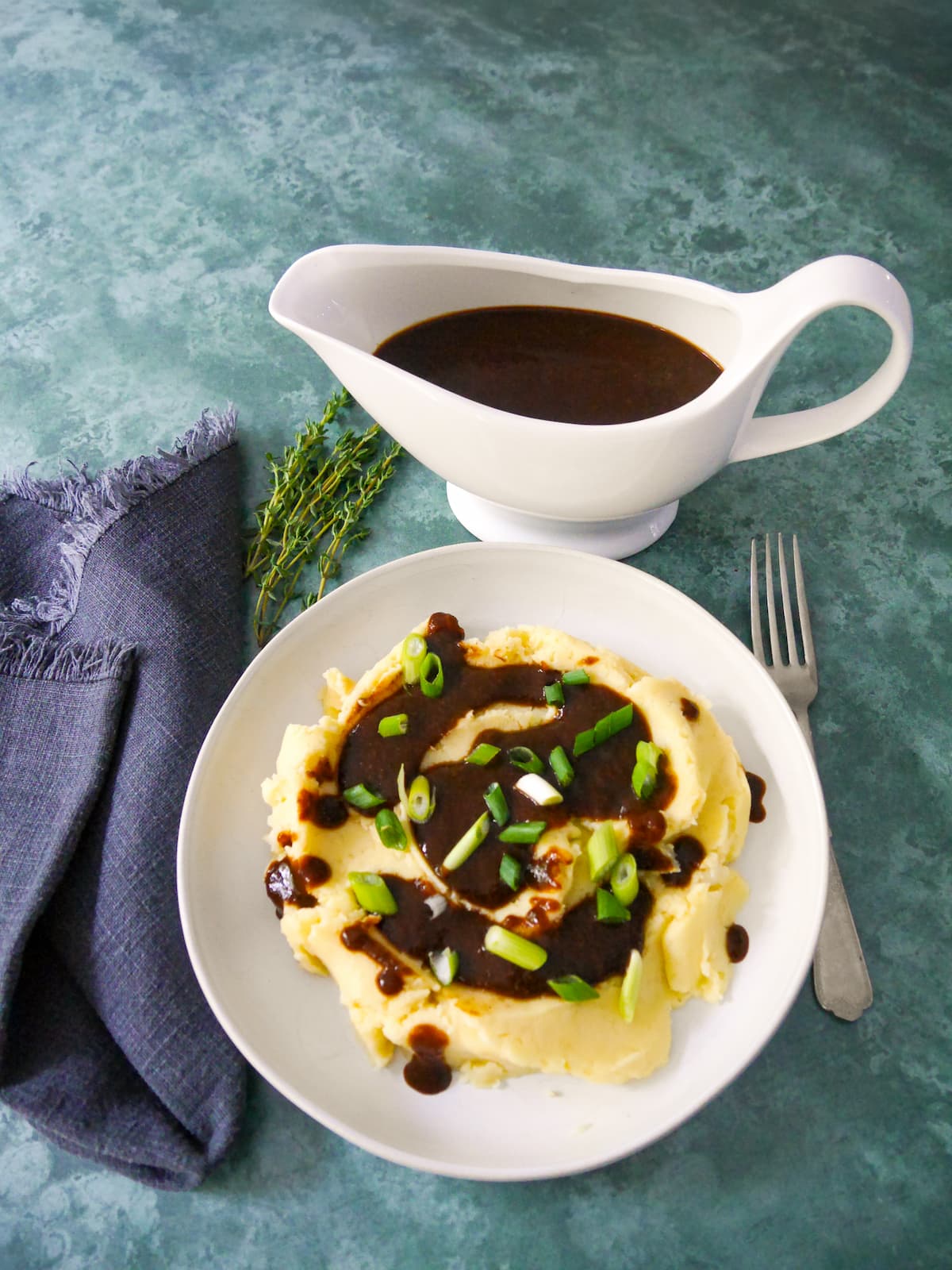 A bowl of creamy mashed potatoes topped with vegan gravy and garnished with sliced green onions, with a gravy boat of gravy set alongside.