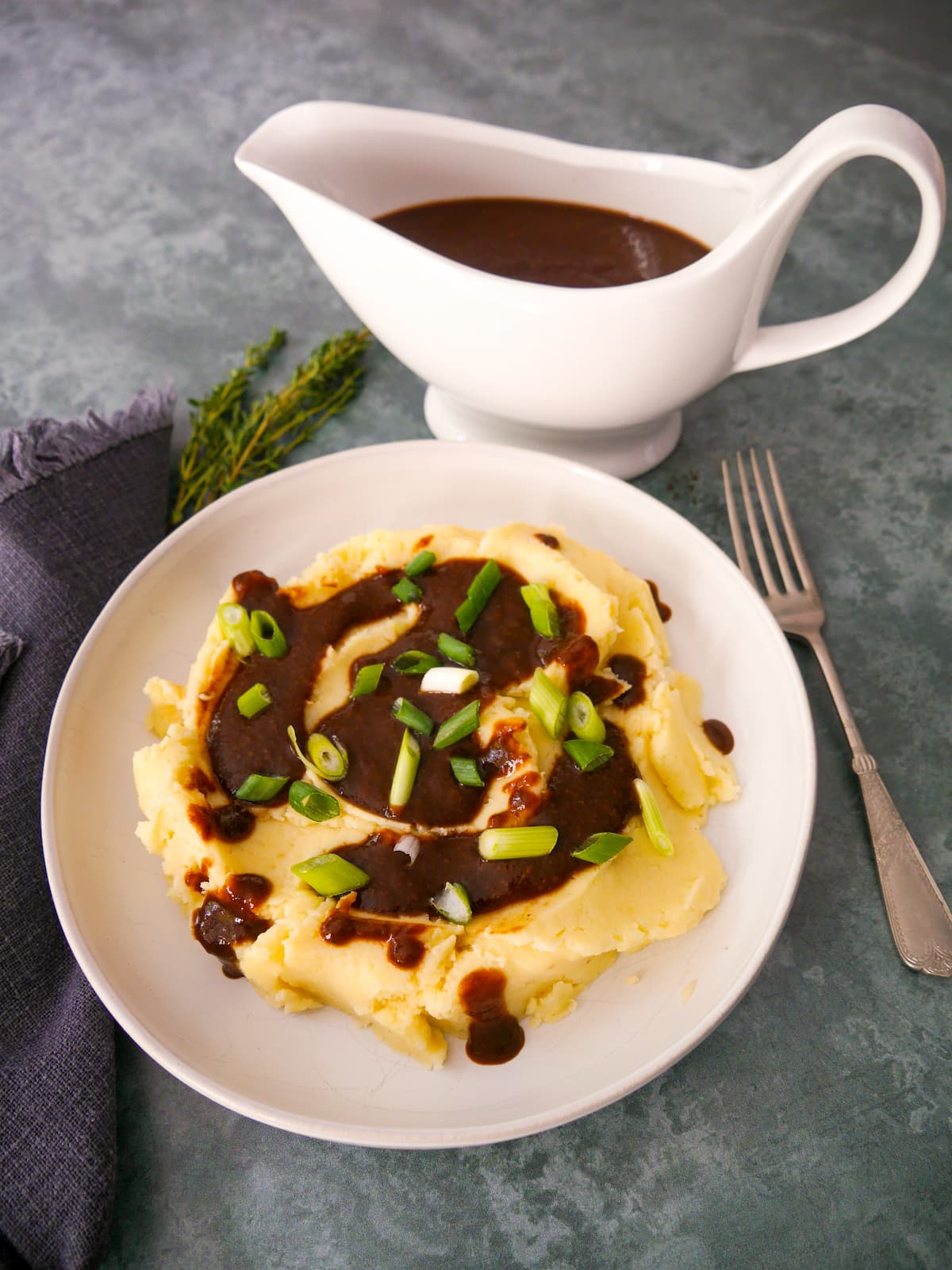 A bowl of creamy mashed potatoes topped with vegan gravy and garnished with sliced green onions, with a gravy boat of gravy set alongside.