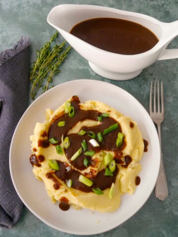 A bowl of creamy mashed potatoes topped with vegan gravy and garnished with sliced green onions, with a gravy boat of gravy set alongside.