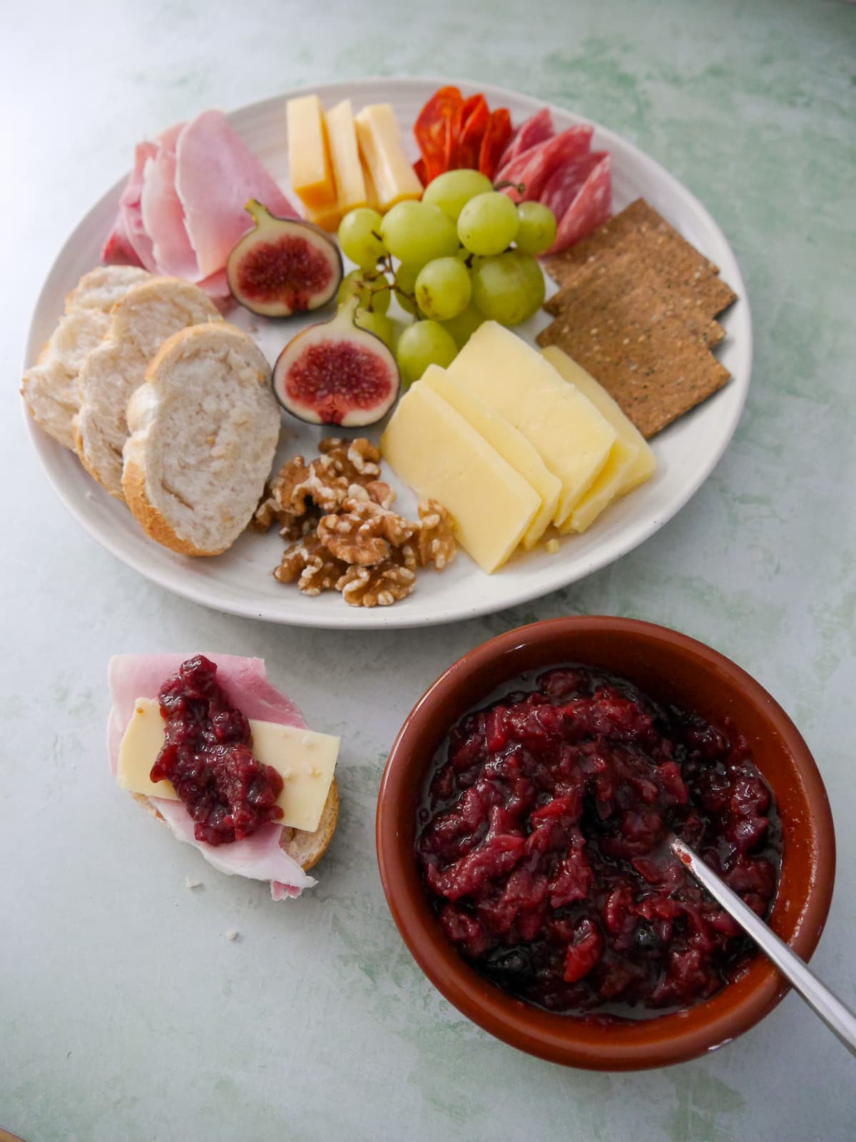 A bowl of apple and cranberry chutney, with a jar of chutney and a plate of sliced bread, crackers, cheese, cold cuts and fruits set alongside.