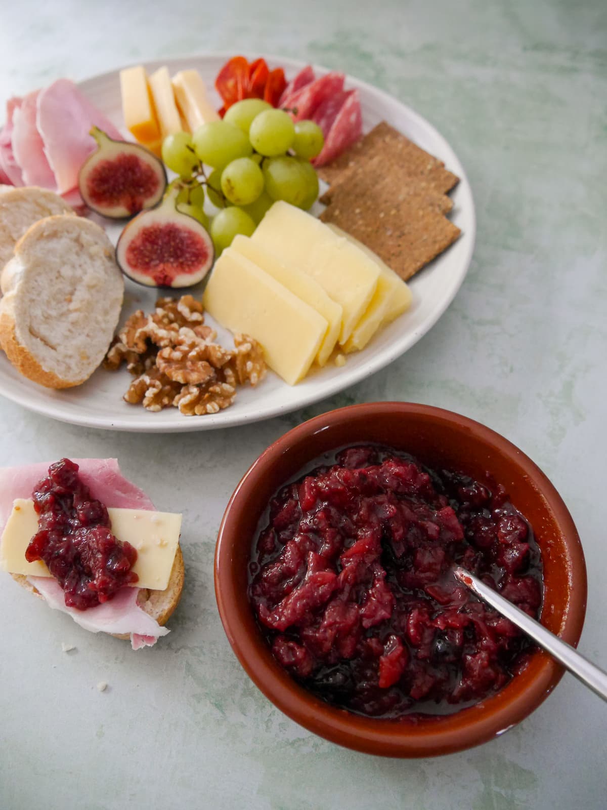 A bowl of apple and cranberry chutney, with a jar of chutney and a plate of sliced bread, crackers, cheese, cold cuts and fruits set alongside.