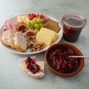A bowl of apple and cranberry chutney, with a jar of chutney and a plate of sliced bread, crackers, cheese, cold cuts and fruits set alongside.