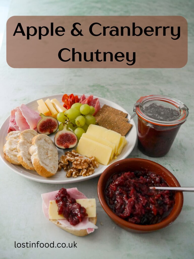 Pinnable image with recipe title and a bowl of apple and cranberry chutney, with a jar of chutney and a plate of sliced bread, crackers, cheese, cold cuts and fruits set alongside.