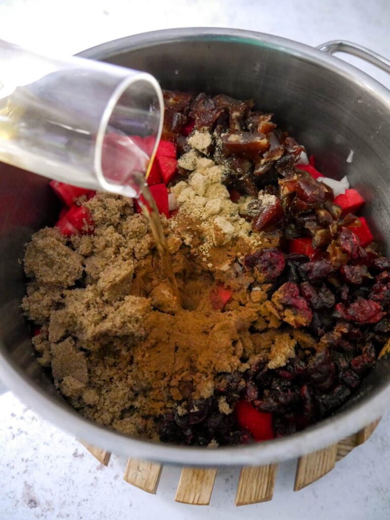 A saucepan of chopped apples, onions, dates and cranberries with added sugar and ground spices with apple cider vinegar being poured over.