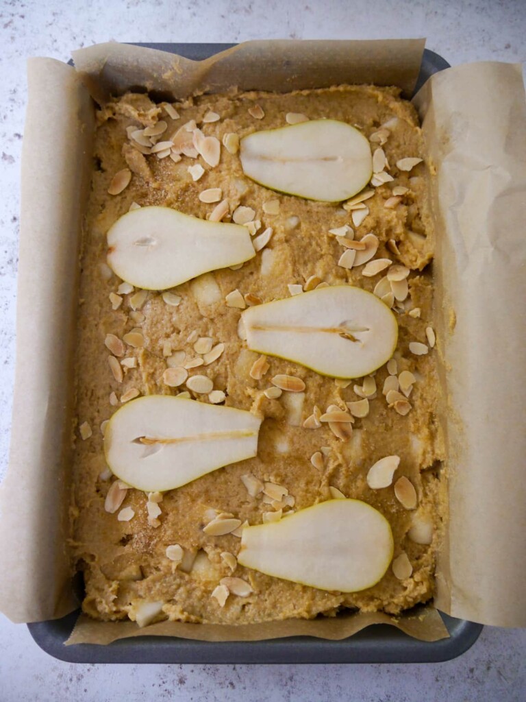 A paper lined baking tray filled with a uncooked pear and almond cake batter, topped with thin slices of pear and flaked almonds.