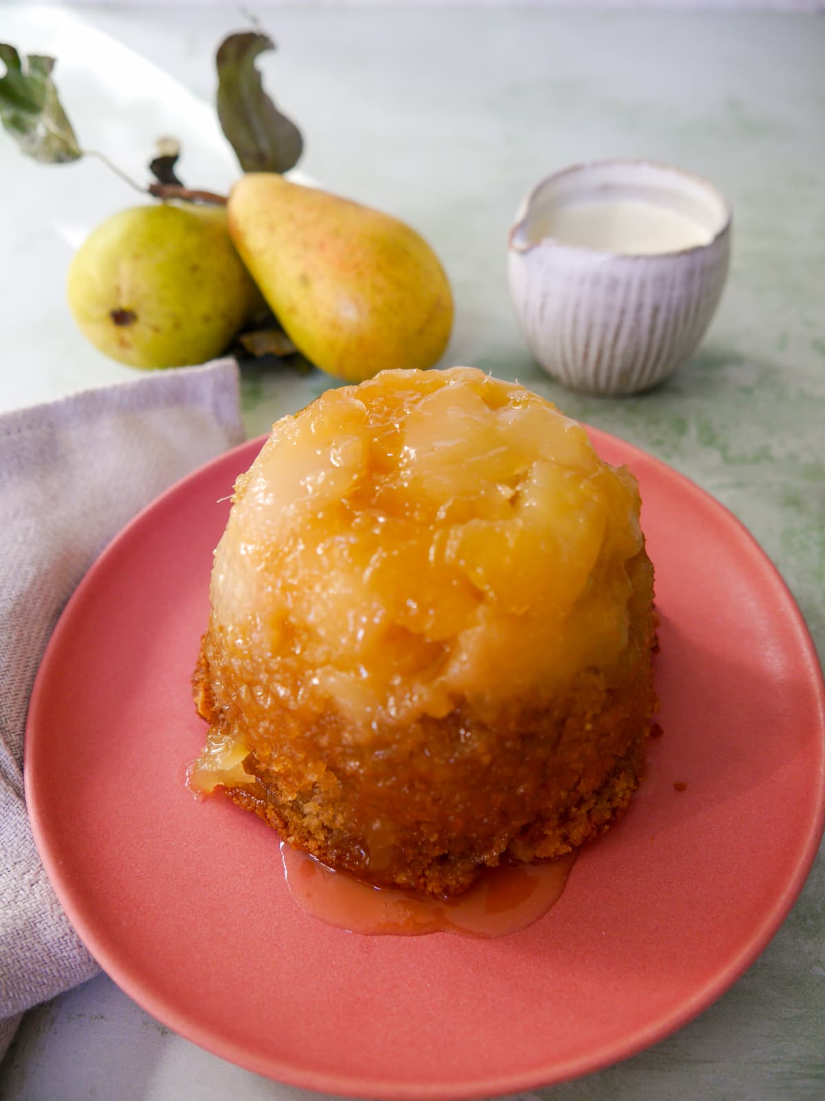 A pink plate topped with and individual pear and ginger sponge pudding with a jug of cream set alongside.