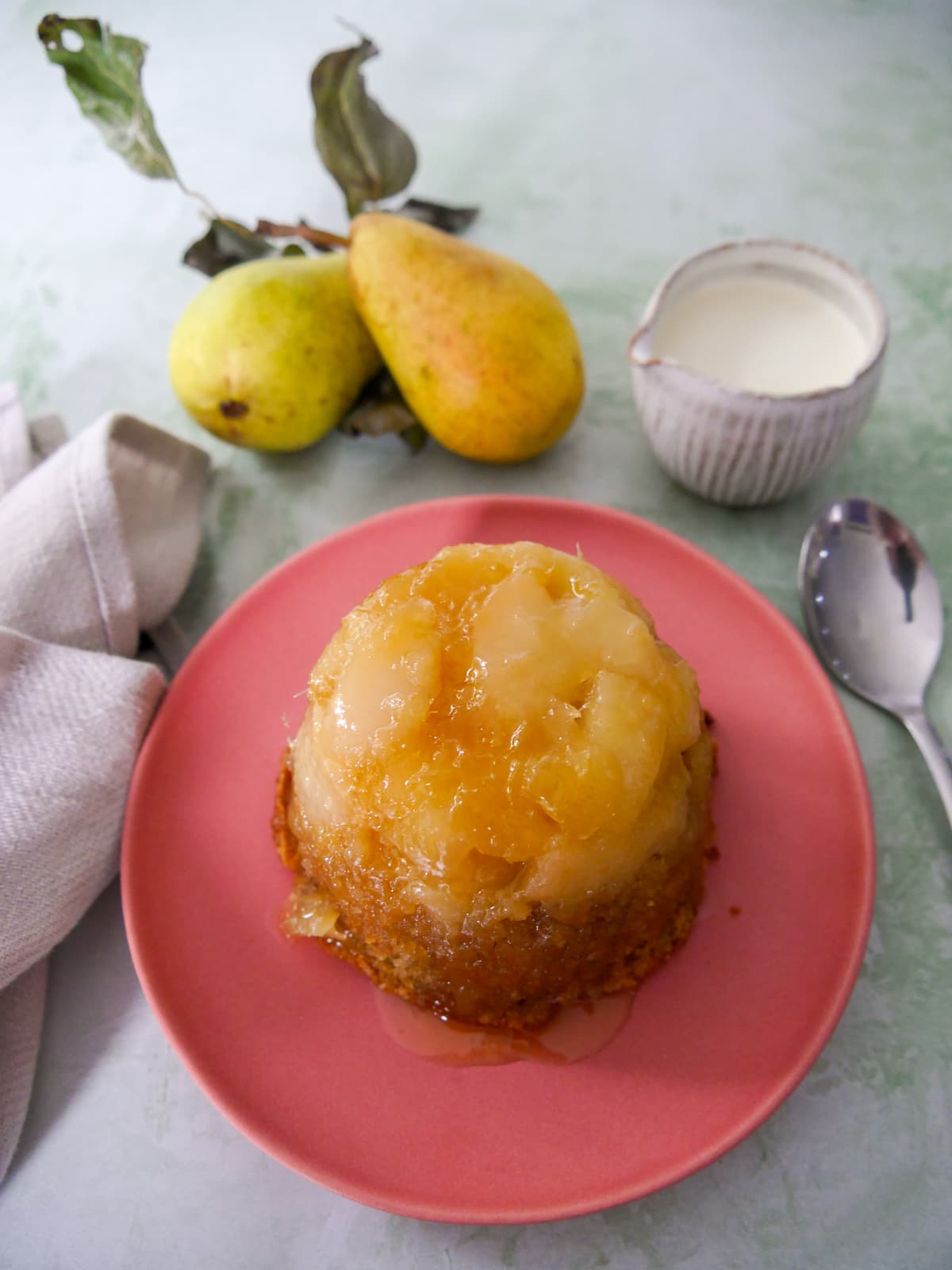 A pink plate topped with and individual pear and ginger sponge pudding with a jug of cream set alongside.