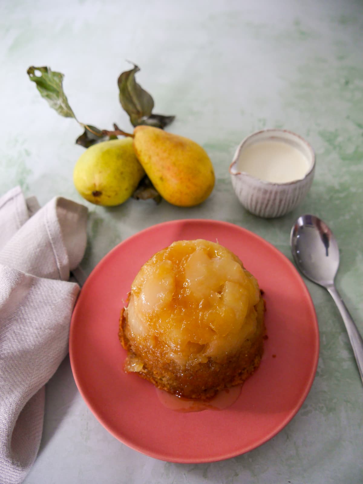 A pink plate topped with and individual pear and ginger sponge pudding with a jug of cream set alongside.