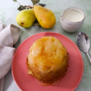 A pink plate topped with and individual pear and ginger sponge pudding with a jug of cream set alongside.