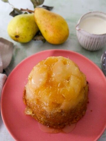 A pink plate topped with and individual pear and ginger sponge pudding with a jug of cream set alongside.
