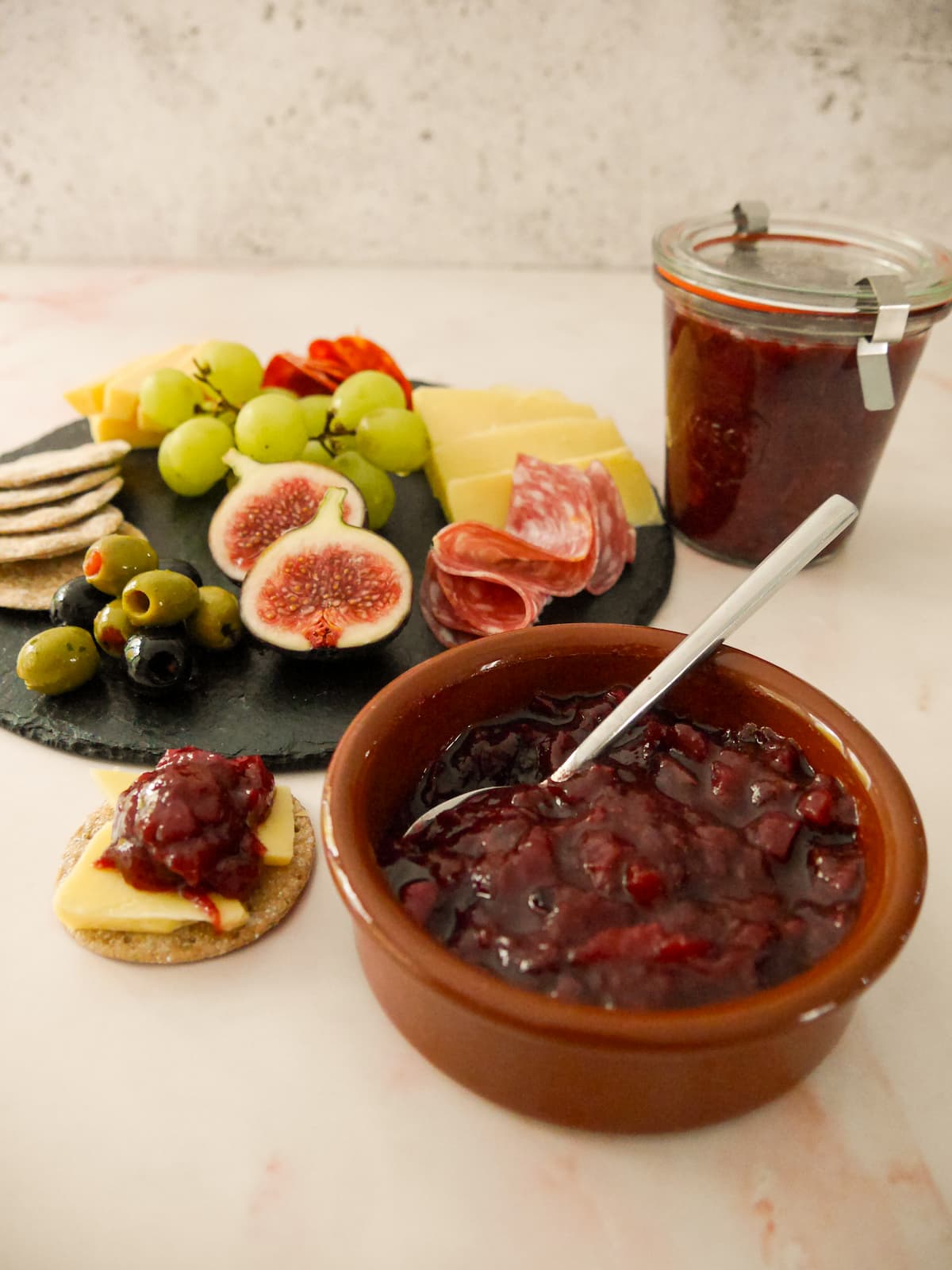 A bowl and jar of spiced plum chutney served with a plate of crackers, cold cuts, olives and fruits set alongside.