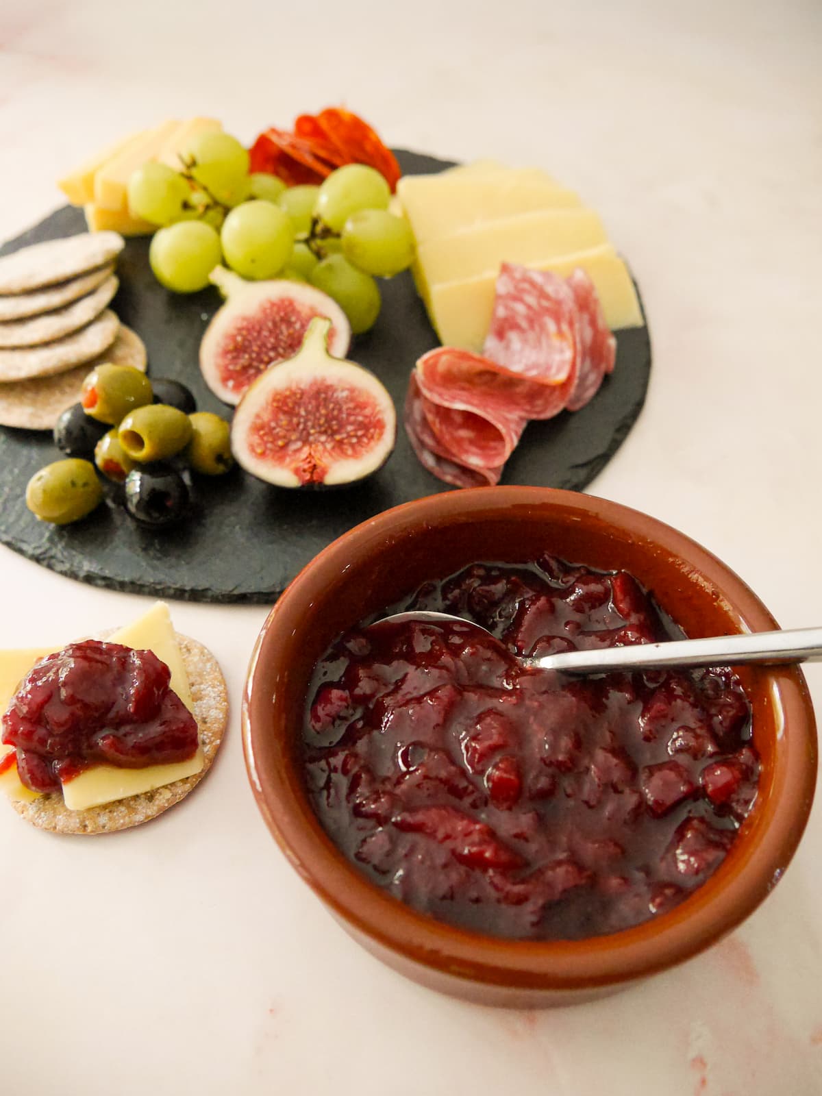 A bowl of spiced plum chutney served with a plate of crackers, cold cuts, olives and fruits set alongside.