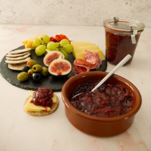 A bowl and jar of spiced plum chutney served with a plate of crackers, cold cuts, olives and fruits set alongside.