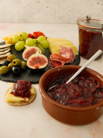A bowl and jar of spiced plum chutney served with a plate of crackers, cold cuts, olives and fruits set alongside.