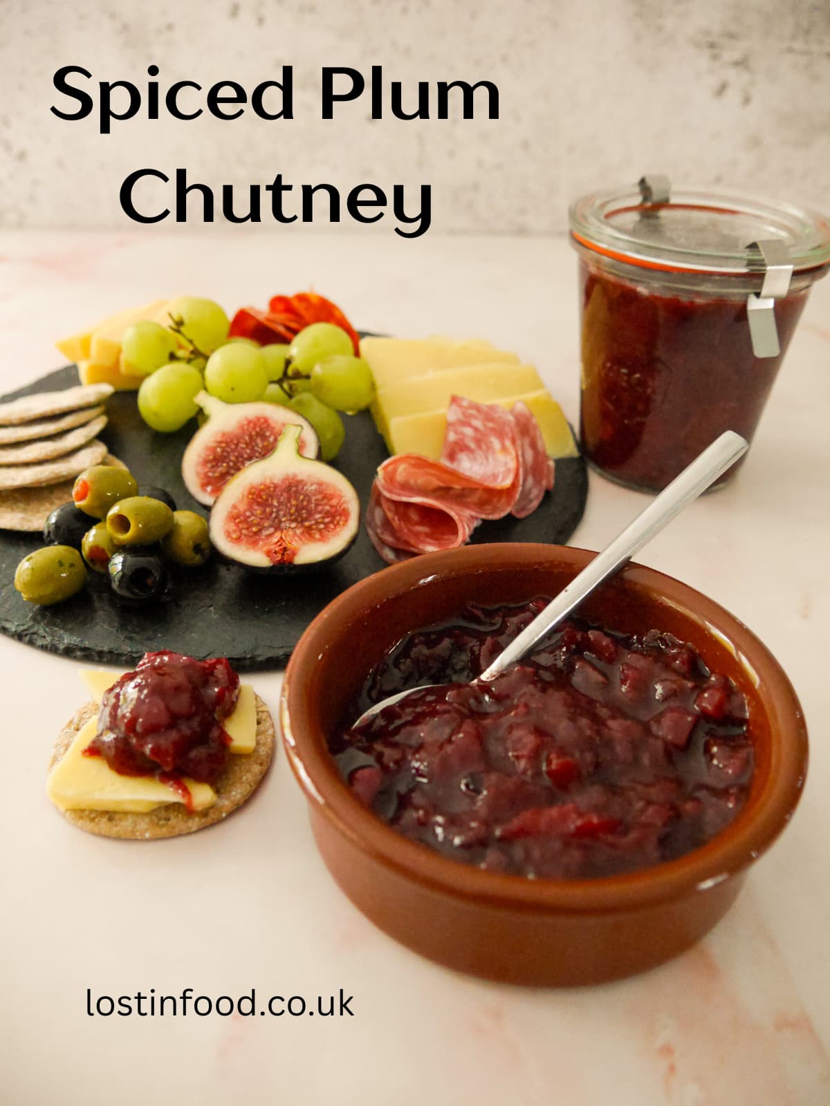 Pinnable image with recipe title and a bowl of spiced plum chutney served with a plate of crackers, cold cuts, olives and fruits set alongside.