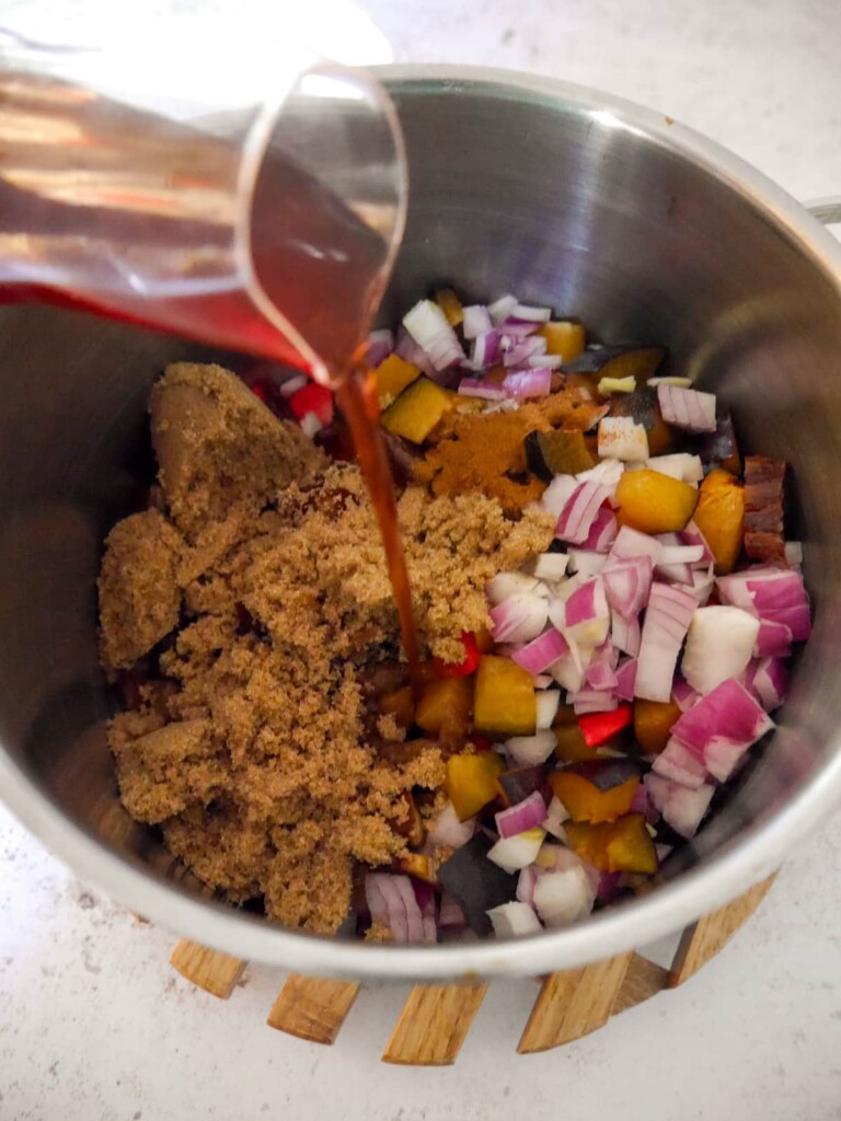 A saucepan filled with chopped mixed vegetables, dates and plums, ground spices and soft brown sugar with red wine being added.