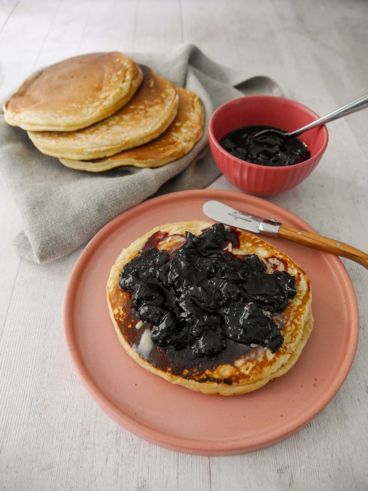 A plate with a pancake topped with black cherry compote, with a bowl of compote and some extra pancakes set alongside.