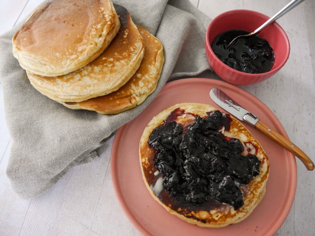 A plate with a pancake topped with black cherry compote, with a bowl of compote and some extra pancakes set alongside.
