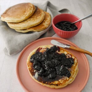 A plate with a pancake topped with black cherry compote, with a bowl of compote and some extra pancakes set alongside.