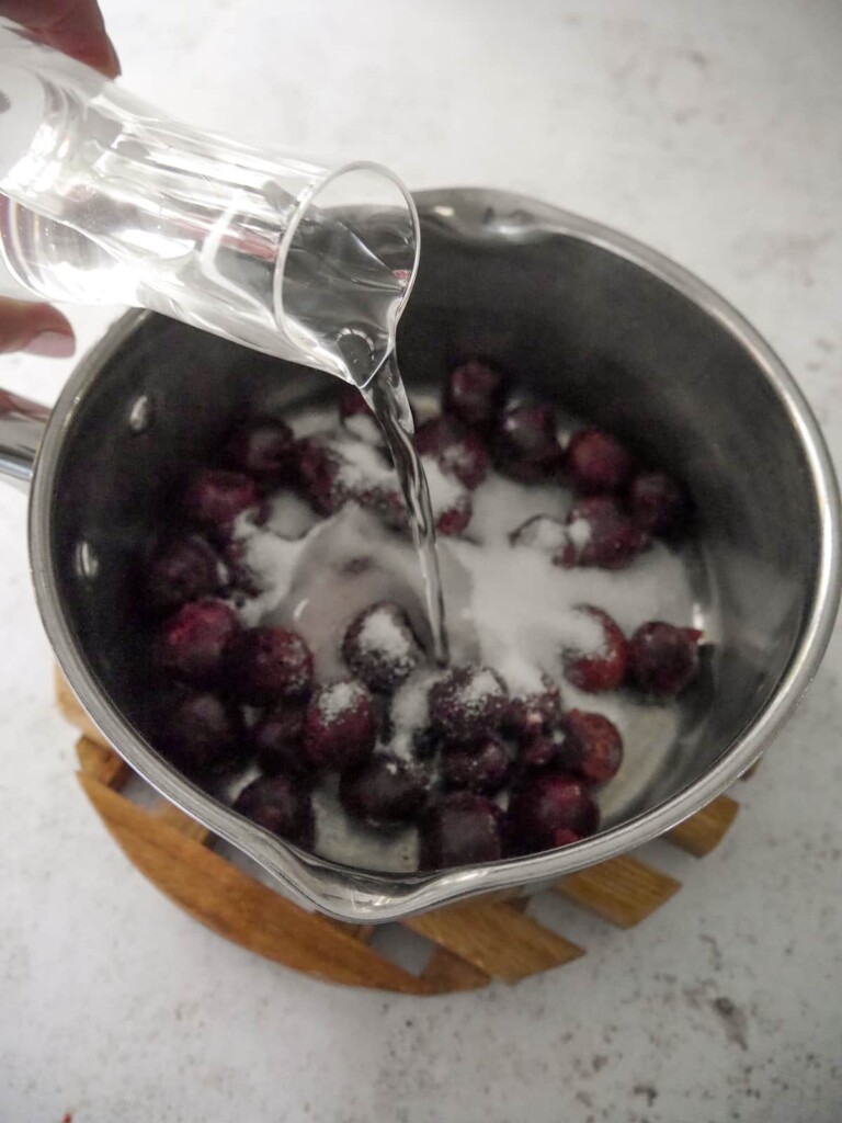 A saucepan of black cherries and sugar with water being added.