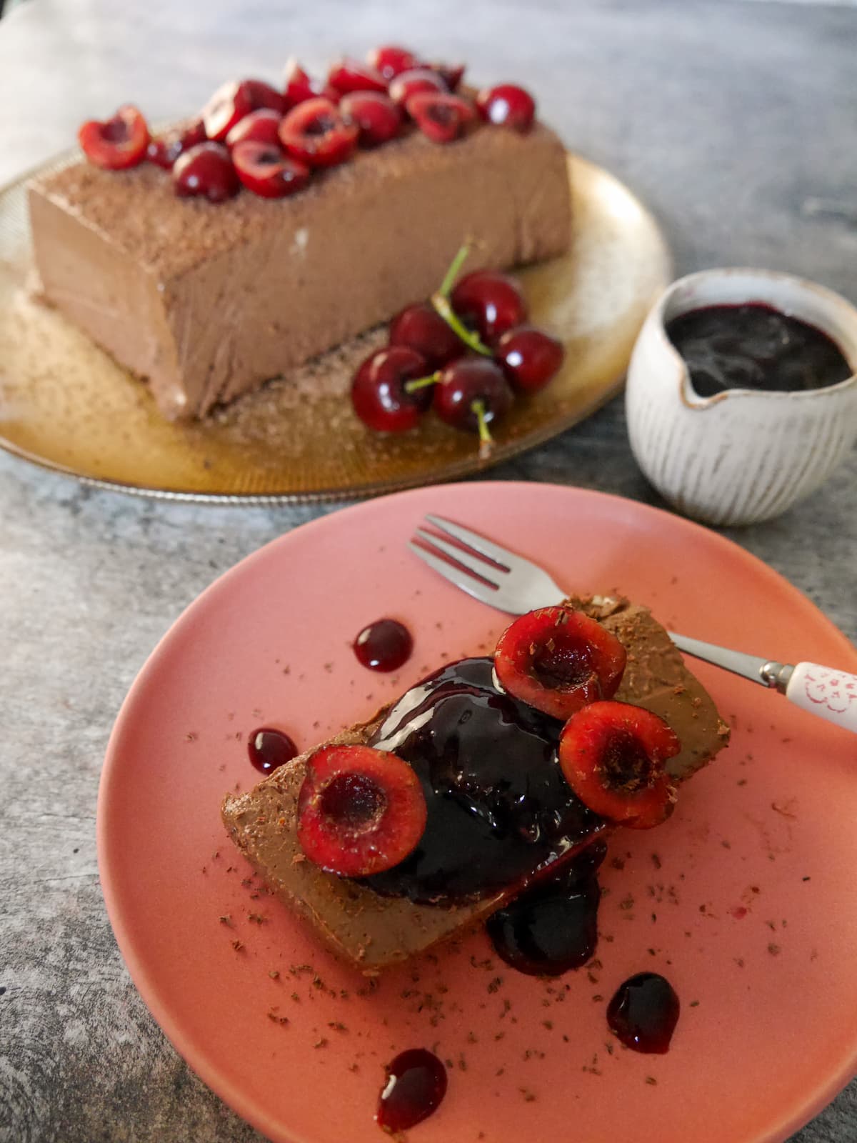 A gold plate topped with frozen dark chocolate parfait, and a plate with a slice of chocolate parfait topped with fresh cherries and cherry compote, and a jug of cherry compote set alongside.