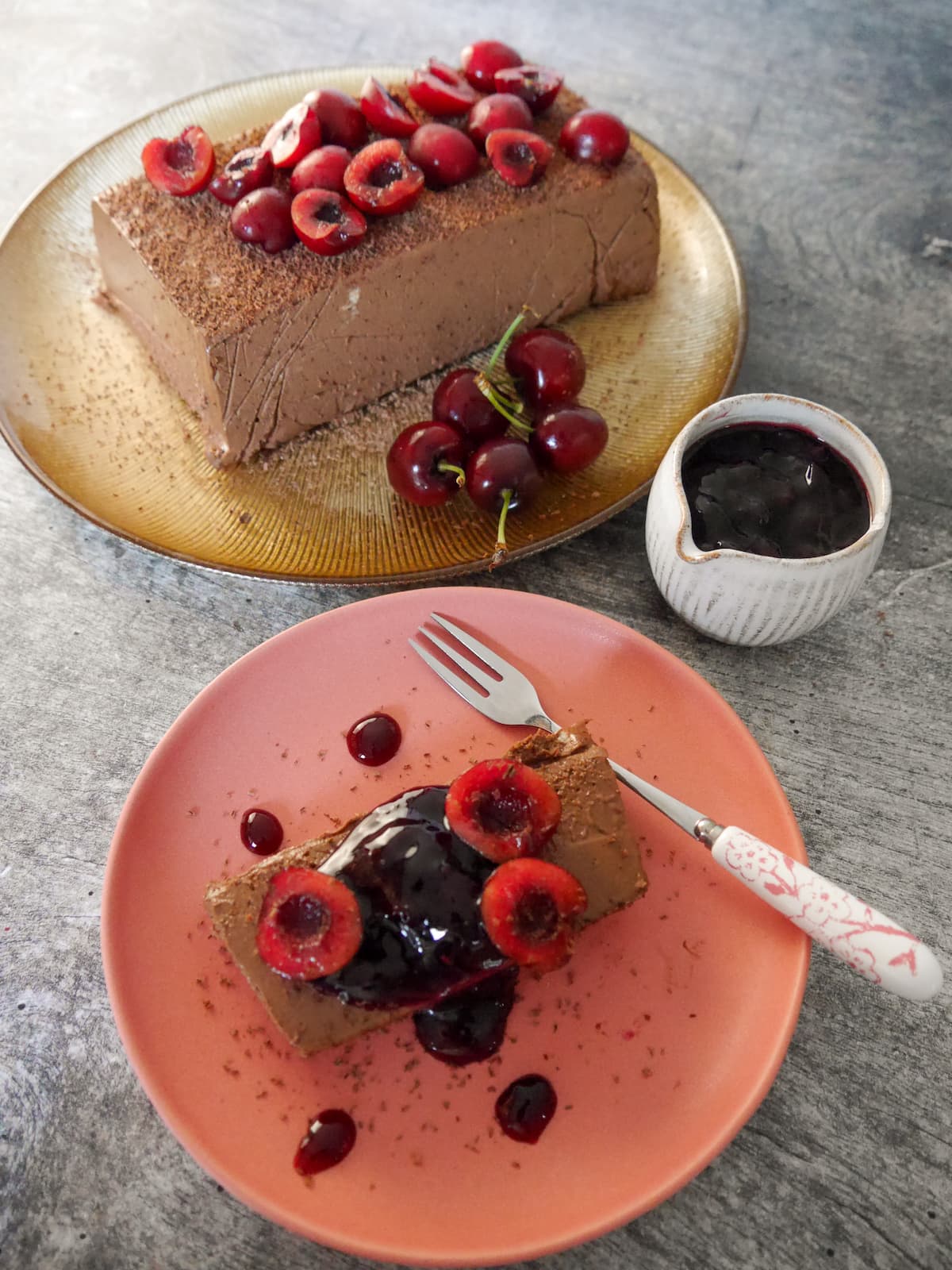 A gold plate topped with frozen dark chocolate parfait, and a plate with a slice of chocolate parfait topped with fresh cherries and cherry compote, and a jug of cherry compote set alongside.