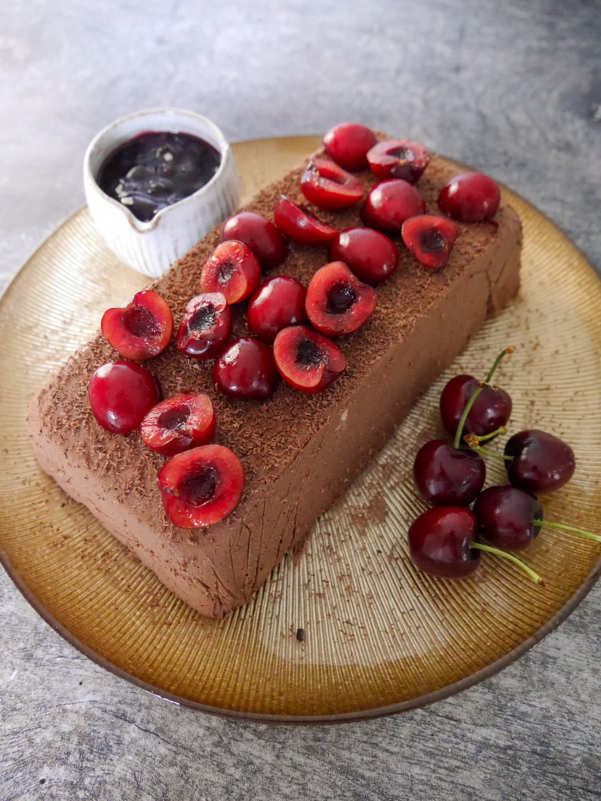 A gold plate topped with frozen dark chocolate parfait, topped with halved fresh cherries and grated 85% dark chocolate, with a jug of dark cherry compote served alongside.