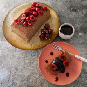 A gold plate topped with frozen dark chocolate parfait, and a plate with a slice of chocolate parfait topped with fresh cherries and cherry compote, and a jug of cherry compote set alongside.