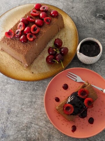 A gold plate topped with frozen dark chocolate parfait, and a plate with a slice of chocolate parfait topped with fresh cherries and cherry compote, and a jug of cherry compote set alongside.