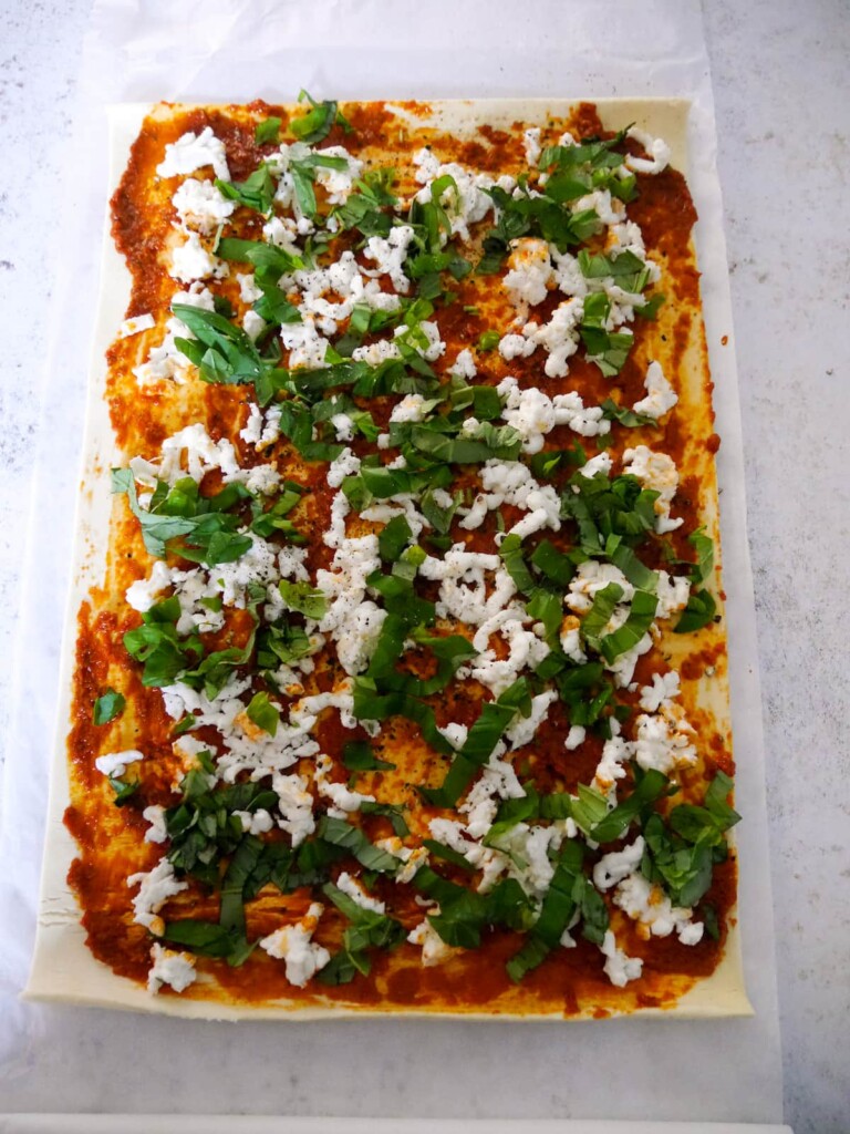 A sheet of puff pastry topped with red pesto, grated fresh mozzarella cheese and shredded basil.