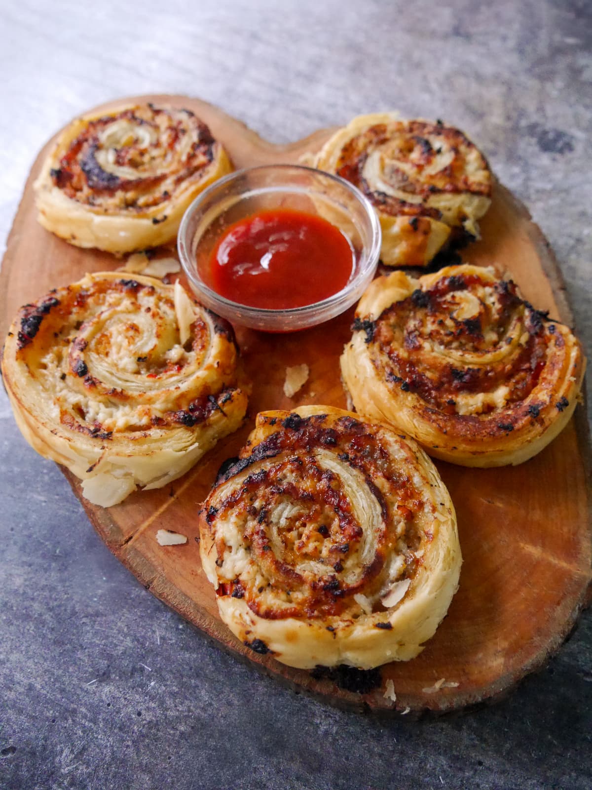 A wooden board topped with puff pastry sausage pinwheels served with a bowl of tomato ketchup.