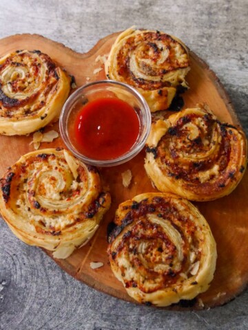 A wooden board topped with puff pastry sausage pinwheels served with a bowl of tomato ketchup.