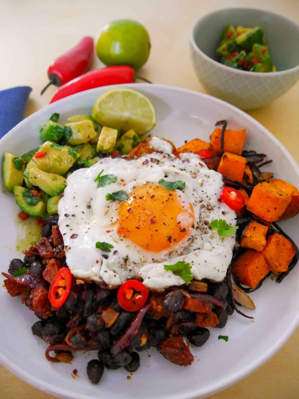 A bowl of cooked chorizo and black beans, roasted sweet potato and avocado salsa topped with a fried egg.