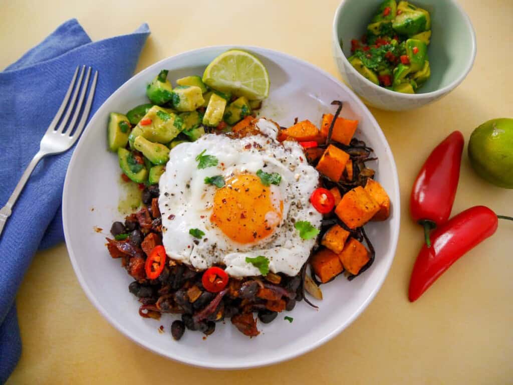 A bowl of cooked chorizo and black beans, roasted sweet potato and avocado salsa topped with a fried egg.