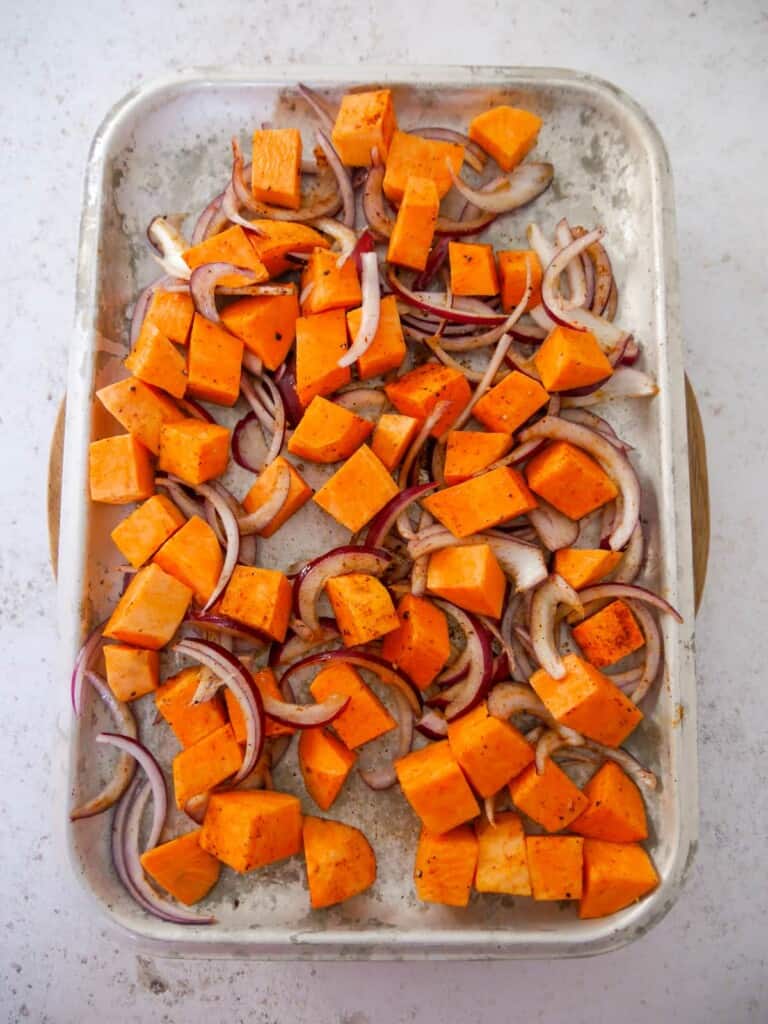 A baking sheet topped with raw sweet potato cubes and sliced red onion, drizzled with olive oil and seasoned with salt and black pepper.