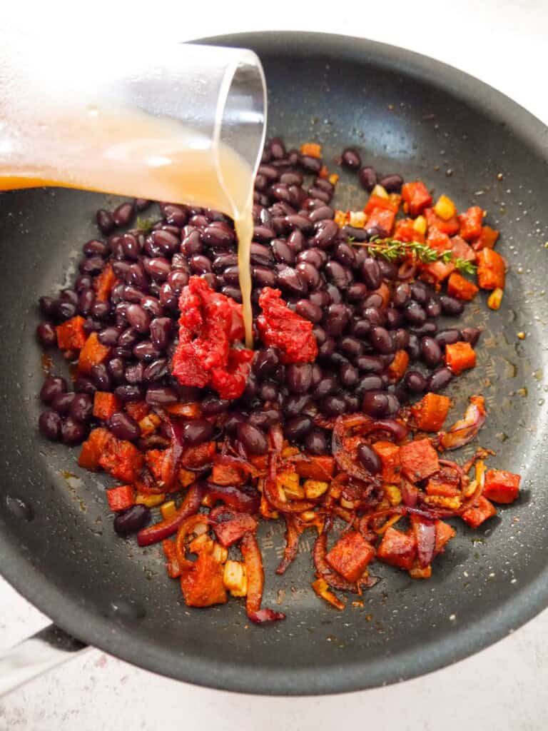 A saucepan of sauteed chorizo, onion and garlic with added black beans, tomato puree, thyme sprigs with chicken stock being poured over.
