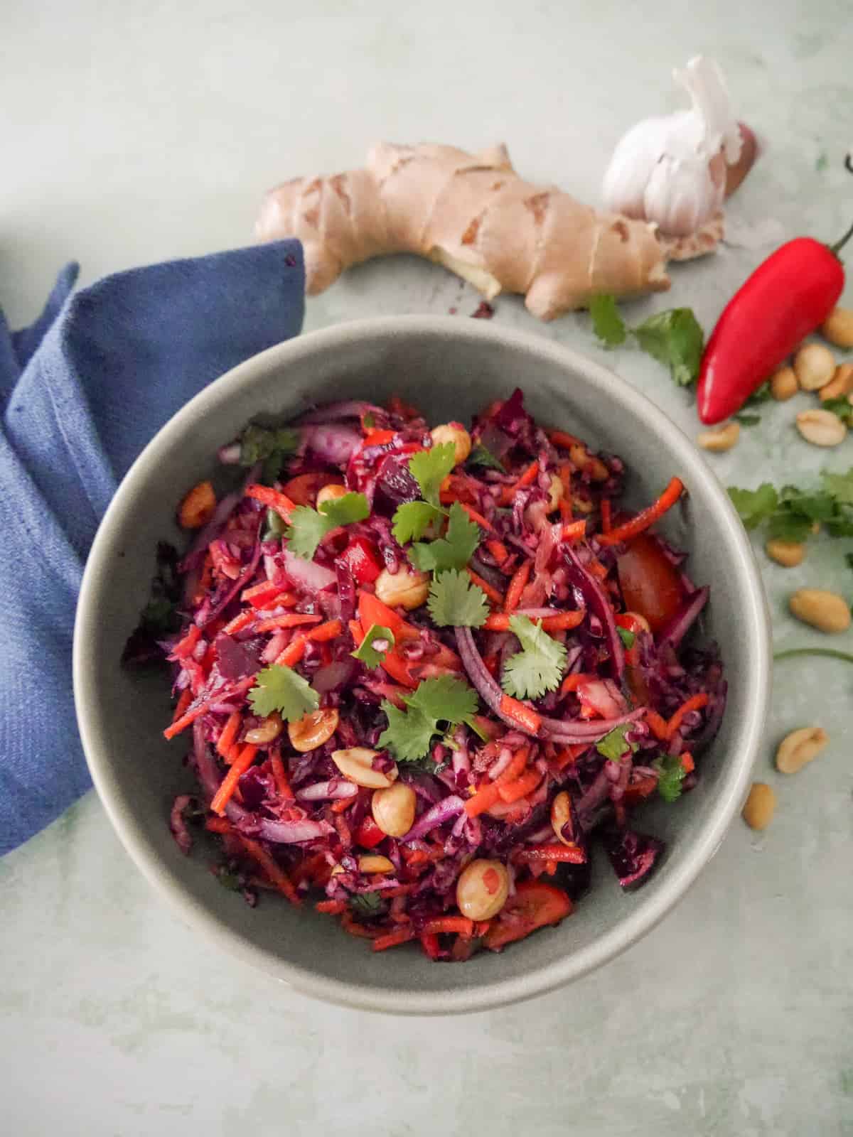 A bowl of roasted peanut and ginger slaw garnished with coriander leaf, with fresh ginger root, a bulb of garlic, red chilli, and peanuts set alongside.