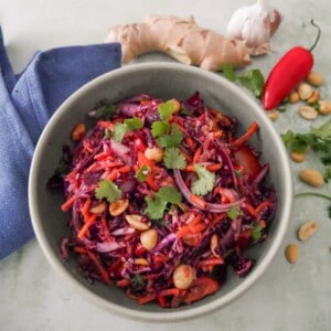 A bowl of roasted peanut and ginger slaw garnished with coriander leaf, with fresh ginger root, a bulb of garlic, red chilli, and peanuts set alongside.