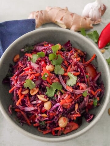 A bowl of roasted peanut and ginger slaw garnished with coriander leaf, with fresh ginger root, a bulb of garlic, red chilli, and peanuts set alongside.