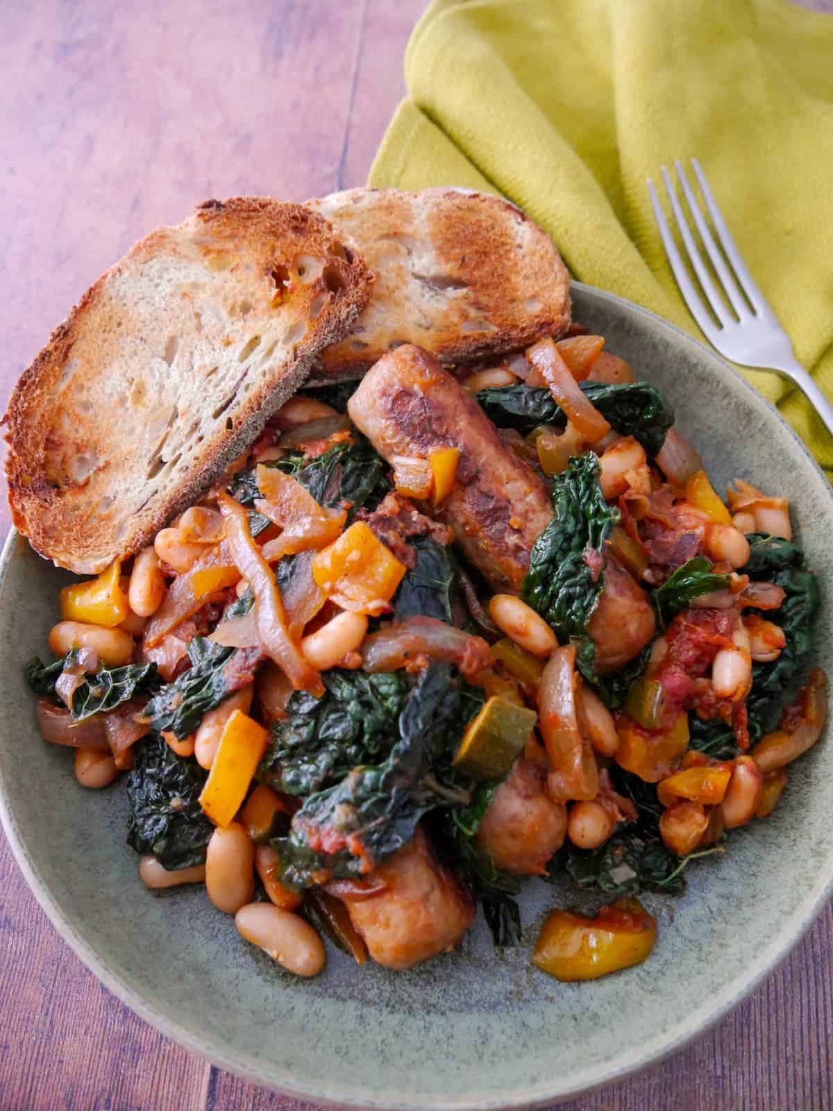 A bowl of sausage casserole with added cannellini beans and cavolo nero kale, served with toasted bread.