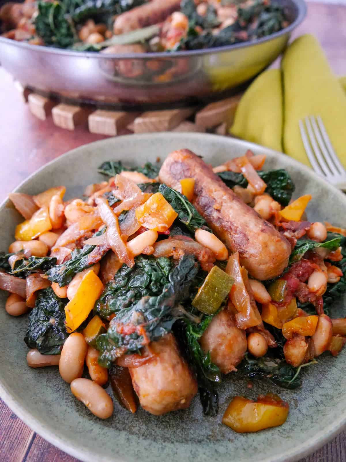 A bowl of sausage casserole with added cannellini beans and cavolo nero kale, served with toasted bread, with a pan of sausage casserole set alongside.