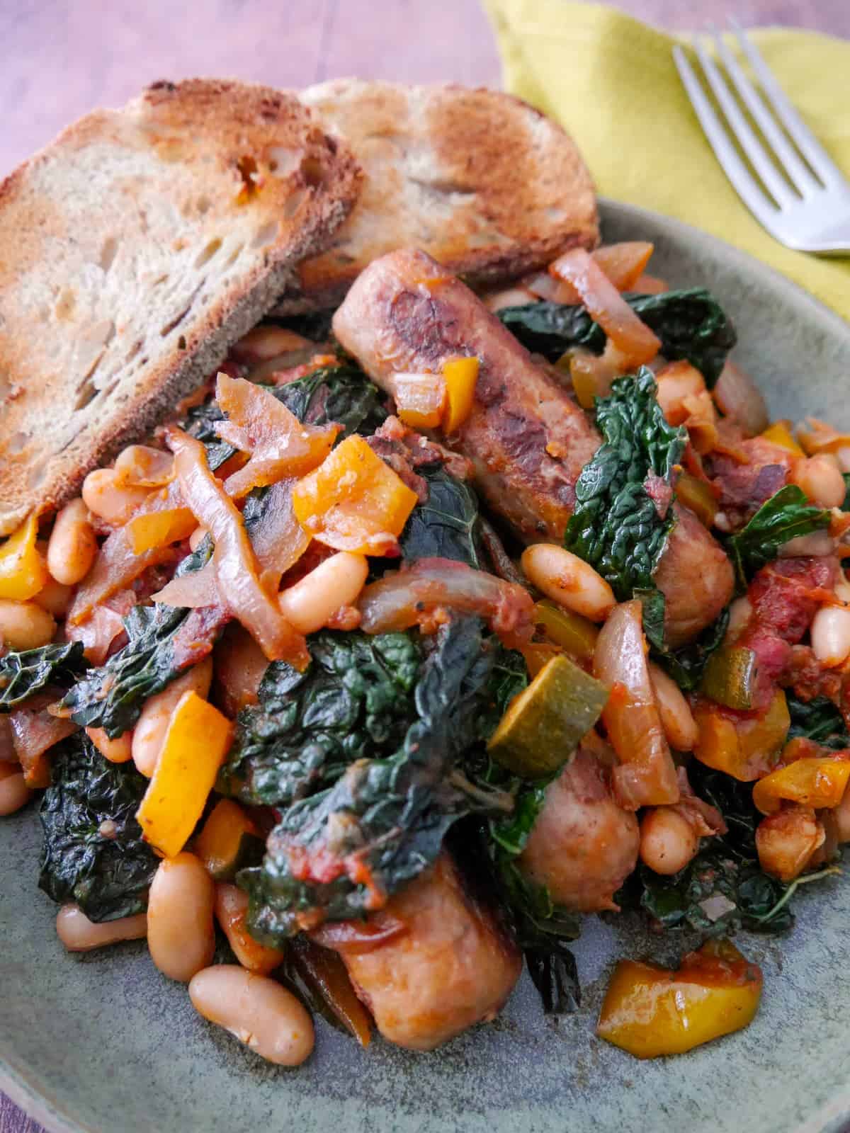 A bowl of sausage casserole with added cannellini beans and cavolo nero kale, served with toasted bread.