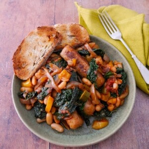 A bowl of sausage casserole with added cannellini beans and cavolo nero kale, served with toasted bread.
