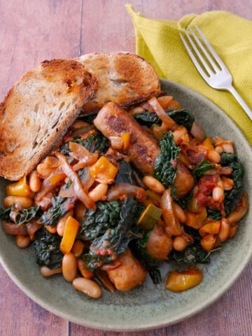 A bowl of sausage casserole with added cannellini beans and cavolo nero kale, served with toasted bread.