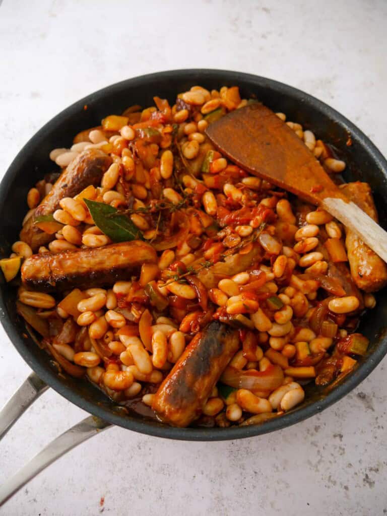 A large saucepan filled with cooked sausages and sauteed vegetables, with added cannellini beans.