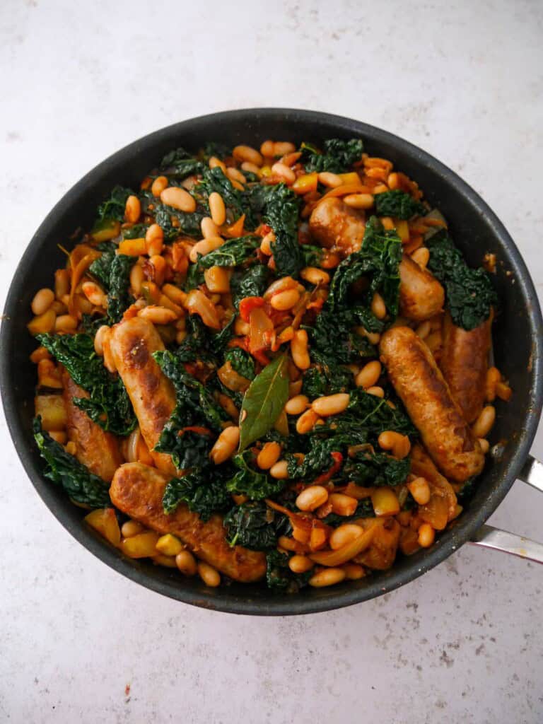 A large saucepan filled with cooked sausages, sauteed vegetables, with cannellini beans and wilted cavolo nero kale.