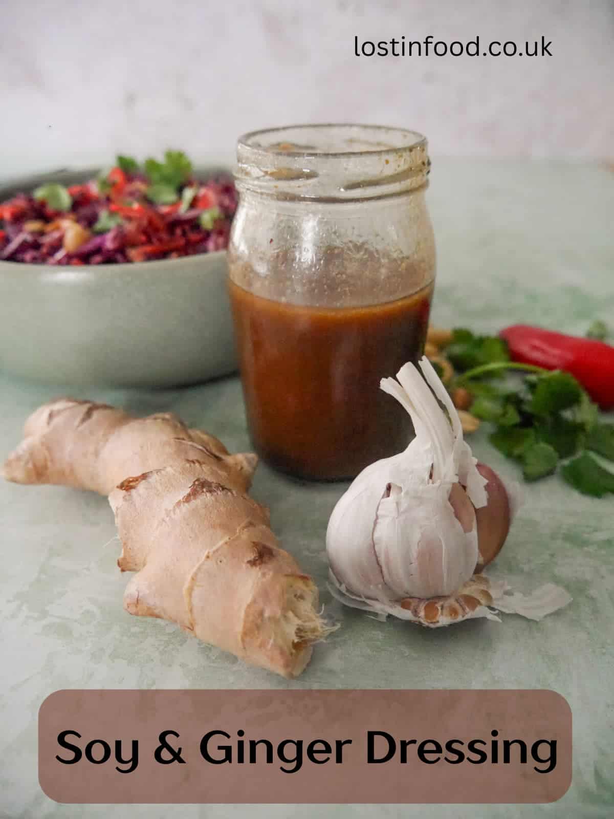 Pinnable image with recipe title and a jar of soy & ginger dressing with fresh root ginger and a bulb of garlic set alongside, and a bowl of peanut and ginger slaw set alongisde.